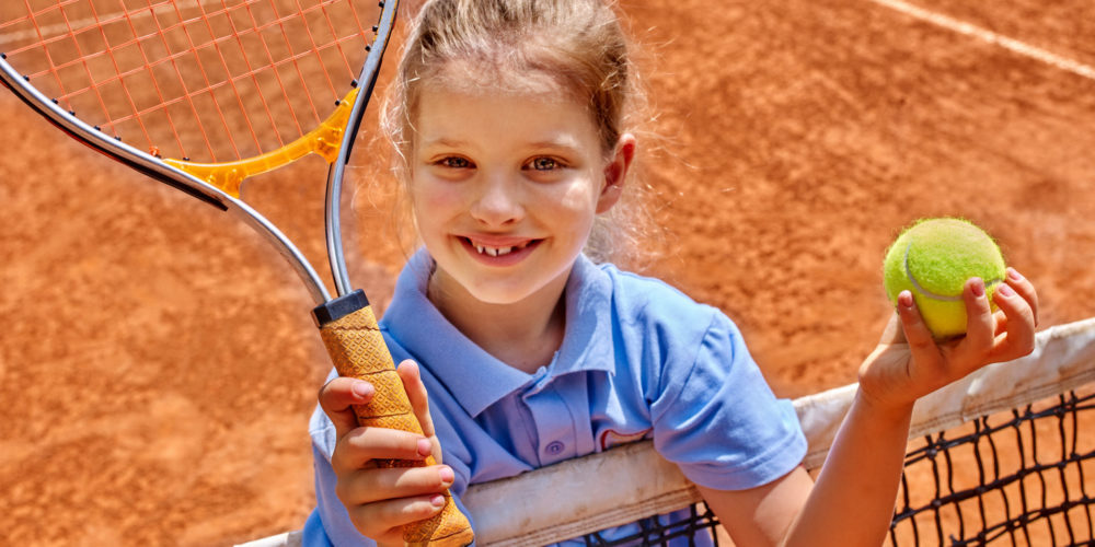Kid athlete in blue form with racket and ball on  brown tennis court.
