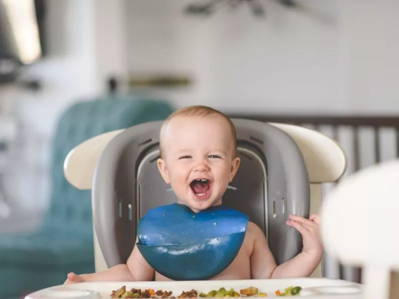 Małe dziecko siedzi na krzesełku do karmienia, śmieje się, je, alergia na pokarm