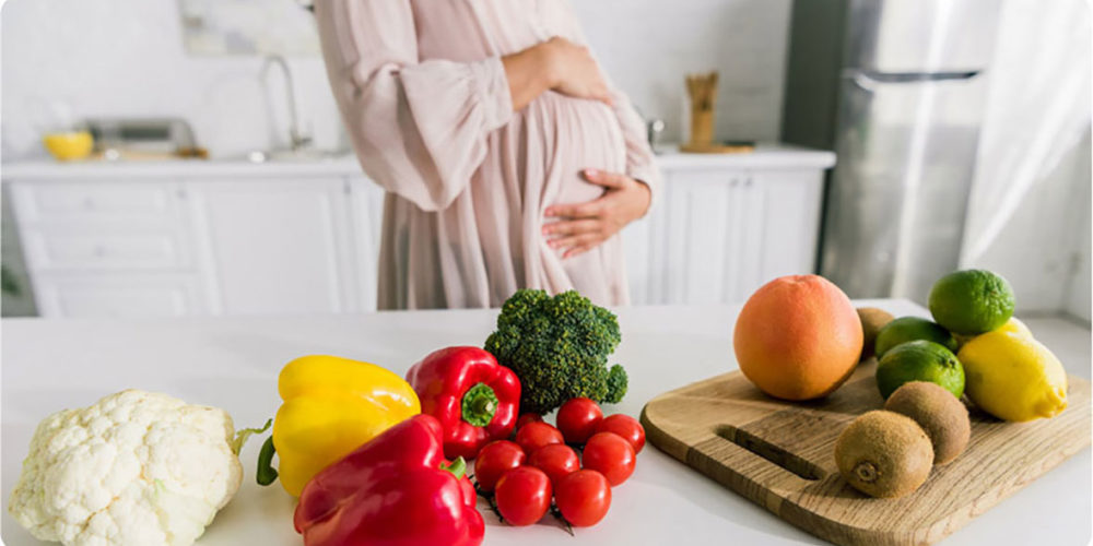 Kobieta w ciąży trzyma się z abrzuch, z przodu leżą owoce i warzywa