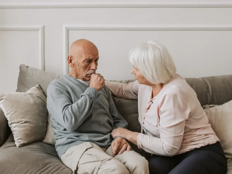 Starszy mężczyzna kaszle na kanapie obok siedzi starsza kobieta, astma