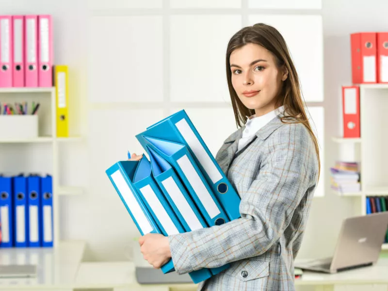 Młoda kobieta w biurze, trzyma w rękach niebieskie segregatory, alergia na pracę